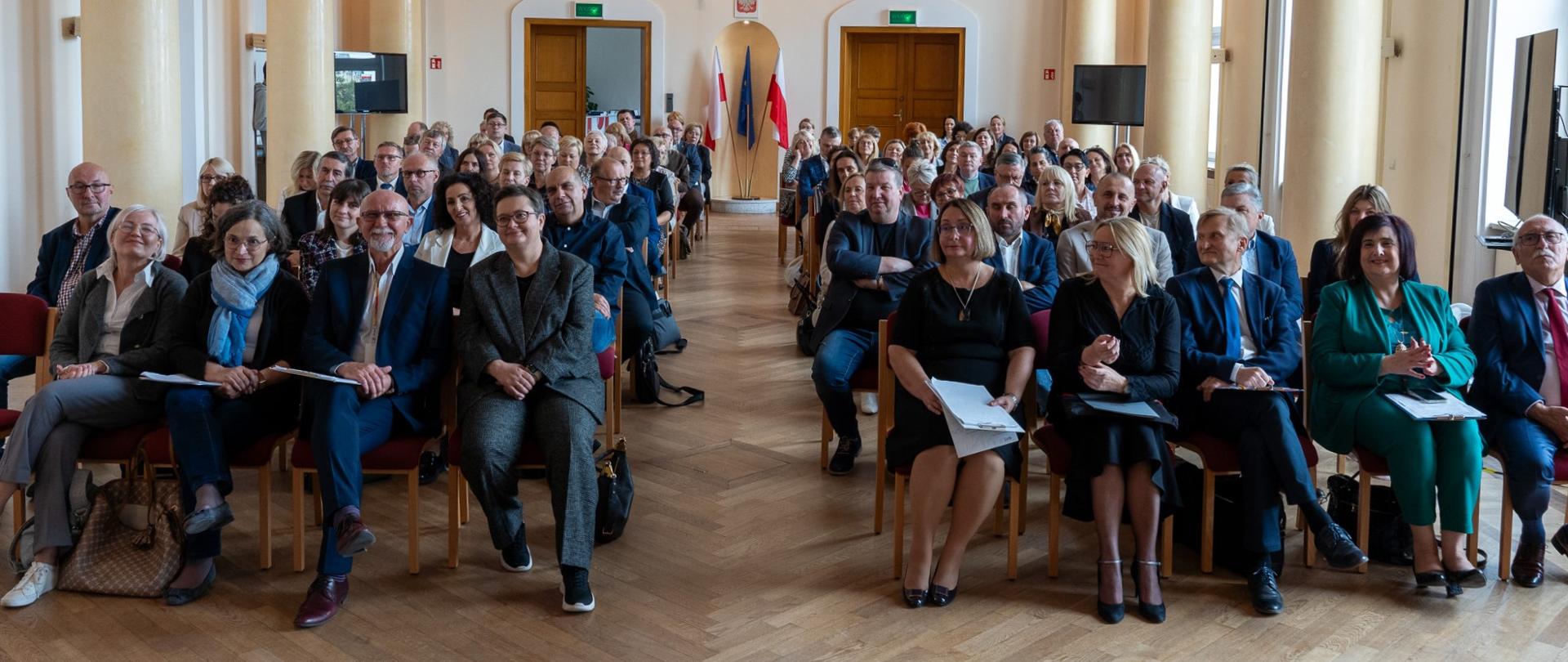 Zdjęcie sali, w której po dwóch stronach ustawione są rzędy krzeseł. Środek pozostaje pusty, umożliwiając swobodne przejście. W pierwszym rzędzie siedzi wiceminister Katarzyna Lubanuer. Sala jest pełna ludzi - uczestników forum. Na ścianie widać napis "docendo discimus". 