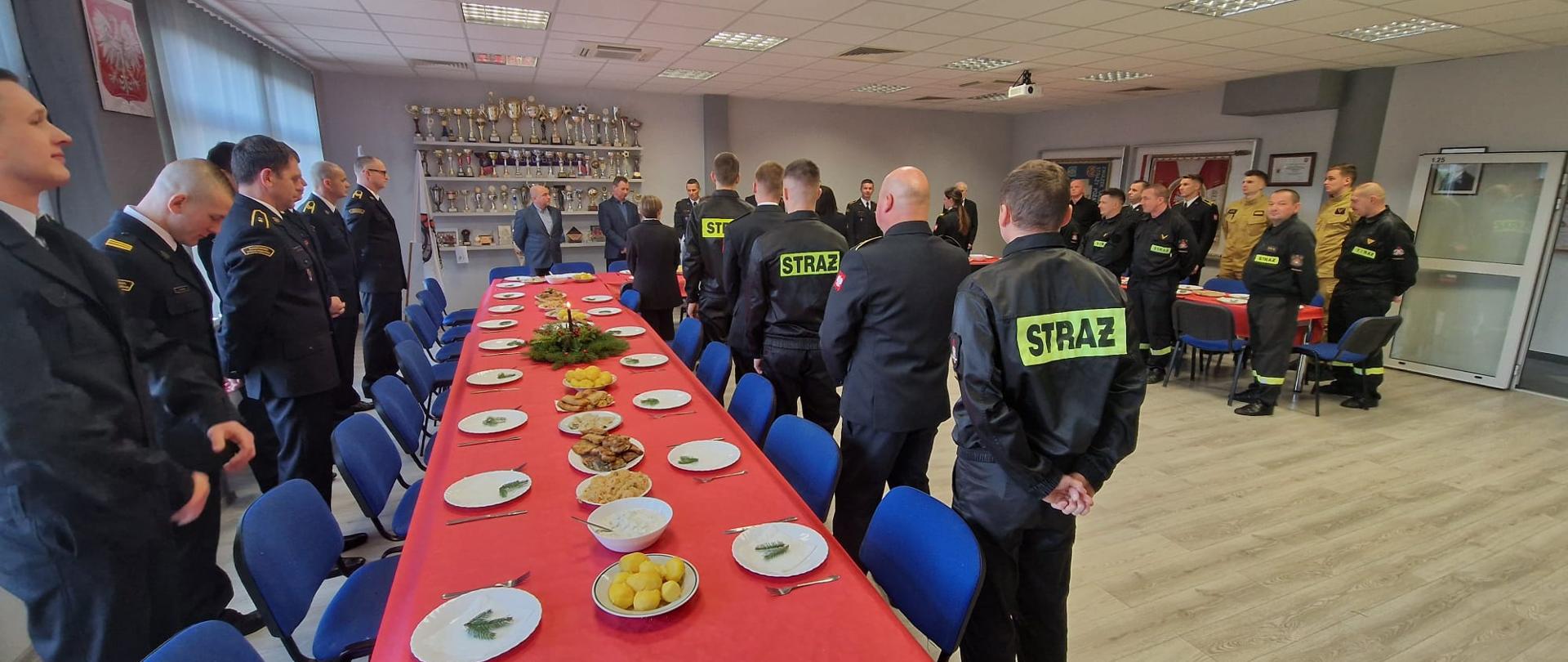 Zdjęcie przedstawia strażaków podczas spotkania wigilijnego. Na zdjęciu widoczne są stoły z potrawami wigilijnymi, stoły nakryte czerwonym obrusem. Wokół stołu stoją strażacy.