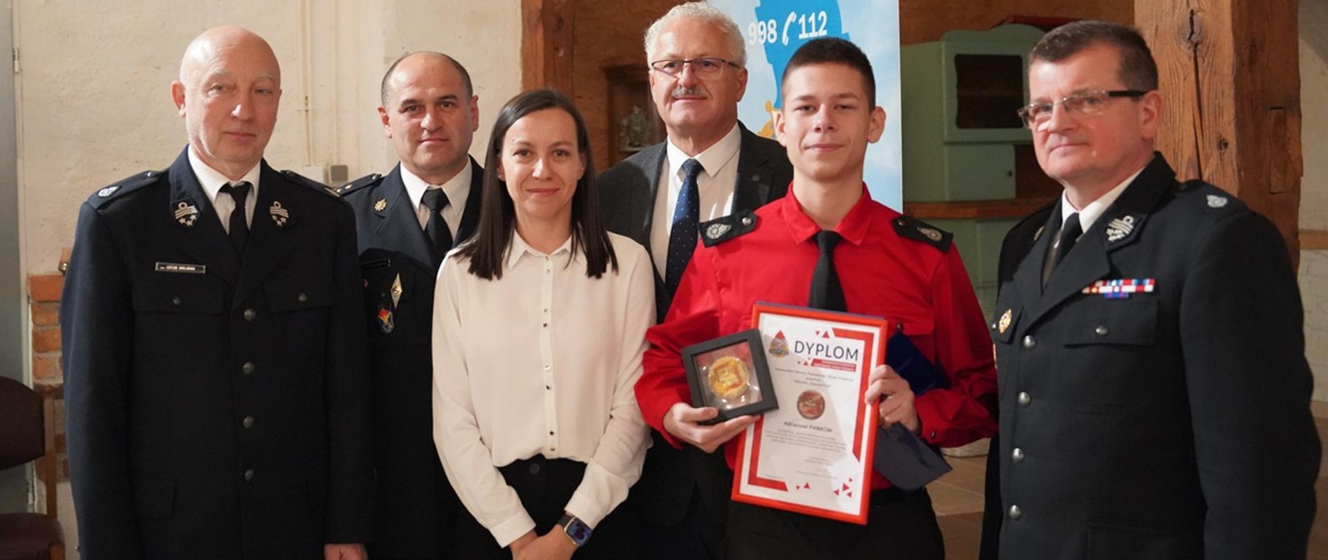 Grupa osób pozuje do zdjęcia. Chłopiec ubrany w czerwoną koszule z czarnym krawatem prezentuje medal oraz dyplom w drewnianej ramce. Obok stoi kobieta w białej koszuli z długimi rękawami. Obok chłopca dwóch druhów w mundurach galowych, funkcjonariusza PSP w mundurze wyjściowym oraz mężczyzna w garniturze.