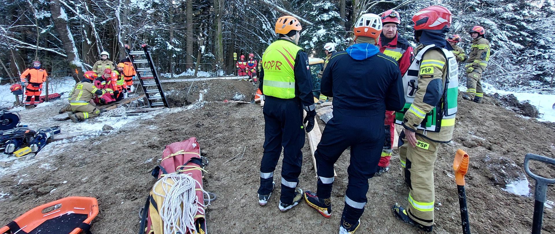 Kolorowa fotografia wykonana na zewnątrz. Przedstawia ratowników różnych służb przy wykopie w którym znajduje się przysypany mężczyzna. Z boku składowany jest sprzęt, ratownicy w ubraniach specjalnych i hełmach, wśród nich Kierujący Działaniem Ratowniczym oznakowany kamizelką.