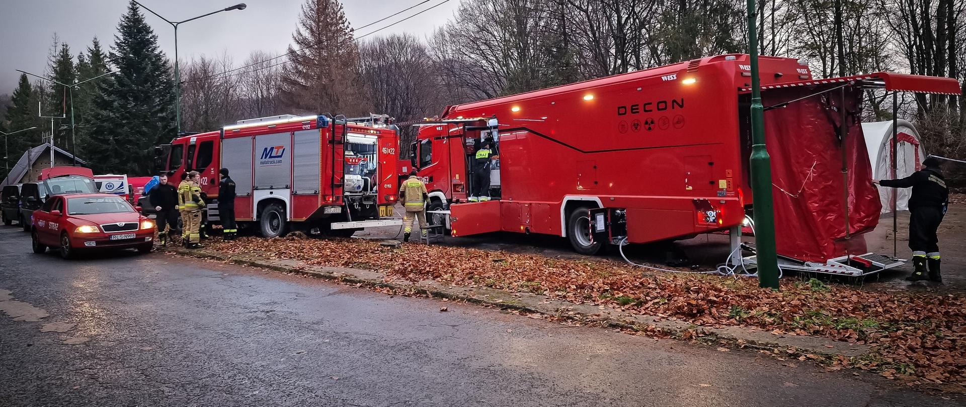 Zdjęcie zrobione na zewnątrz, podczas ćwiczeń. Po prawej stronie stoi Specjalistyczny samochód ratownictwa chemicznego Państwowej Straży Pożarnej. Obok niego strażacy ratownicy ubrani w stroje ochronne.