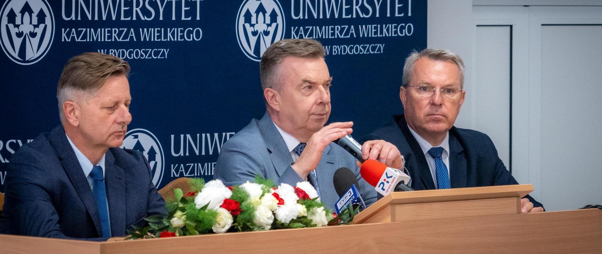 Minister Wieczorek siedzi za stołem, mówi do kilku mikrofonów, obok niego siedzi dwóch mężczyzn w garniturach, za nimi granatowa ścianka z napisem Uniwersytet Kazimierza Wielkiego w Bydgoszczy.