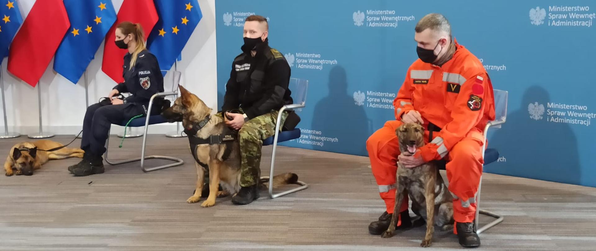 Funkcjonariusz Państwowej Straży Pożarnej, Straży Granicznej oraz Policji siedzą na fotelach, obok nich siedzą psy ratownicze i tropiące. W tle flagi Polski oraz UE.