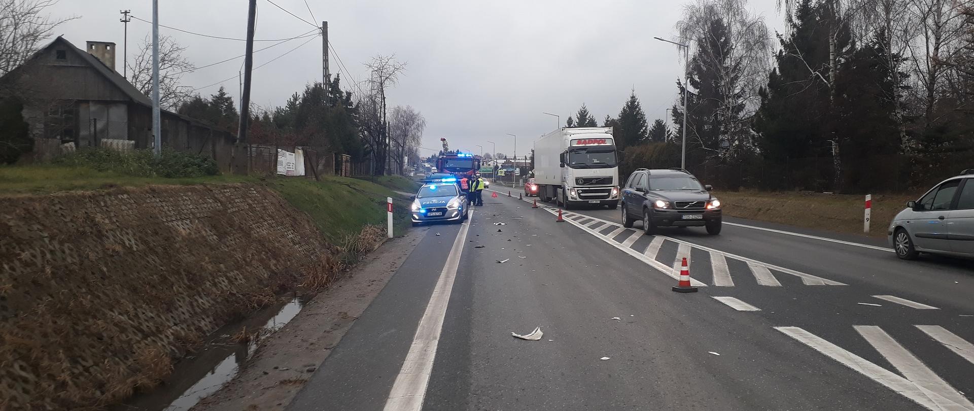 Zdjęcie zrobione w porze dziennej. Widać drogę. Po lewej oznakowany radiowóz policji a po prawej jedzie sznur pojazdów. Na jezdni leża odłamki części samochodowych.