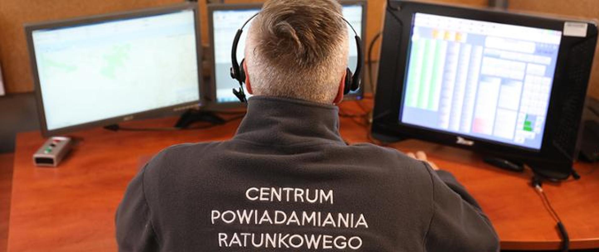 Zdjęcie przedstawia operatora numeru alarmowego 112. Widok na plecy i monitory.