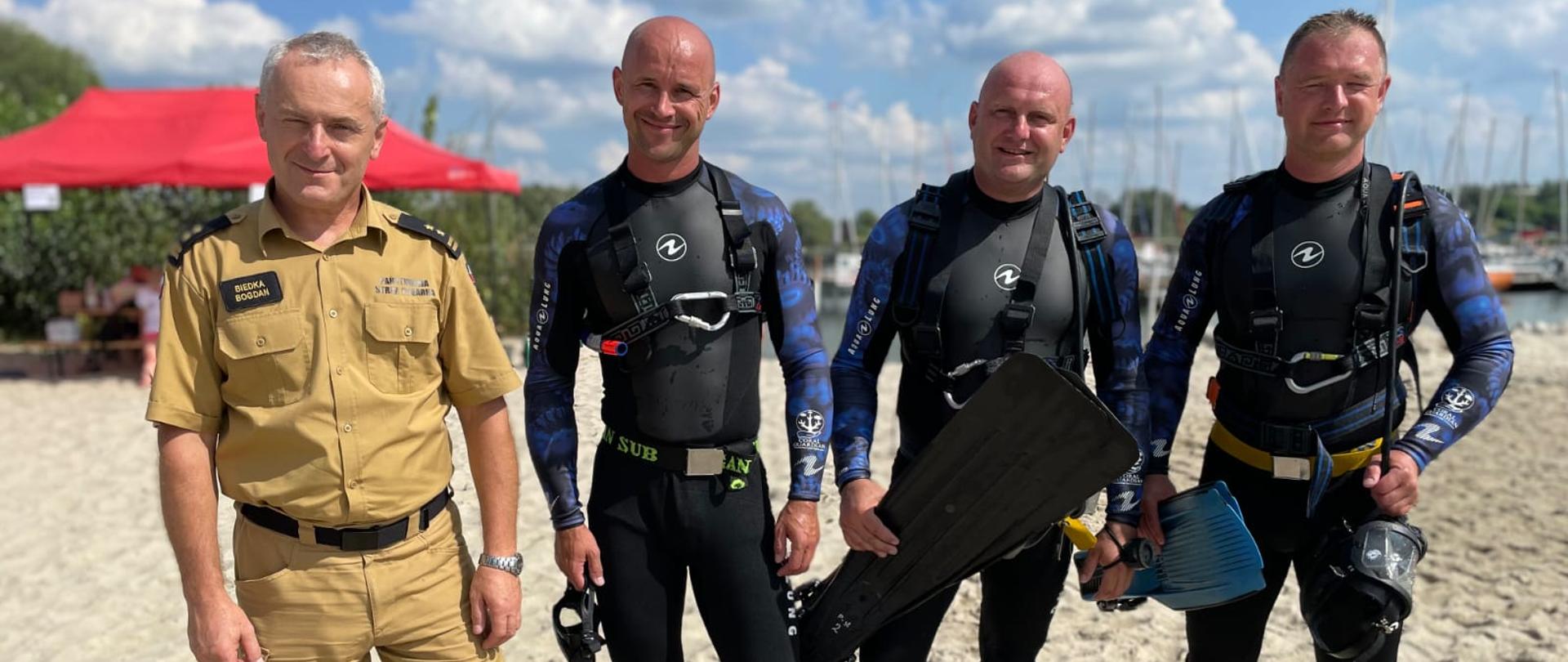 Kolorowa fotografia wykonana na zewnątrz w pogodny dzień na plaży jeziora. Przestawia 3 strażaków płetwonurków ubranych w skafandry nurkowe, jeden ze strażaków ubrany w ubranie służbowe.