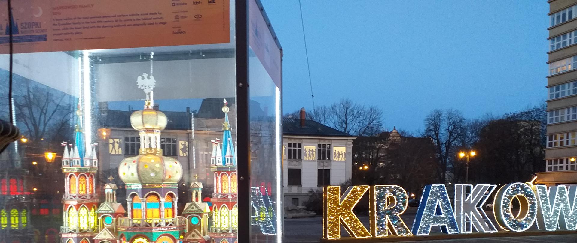 Na zdjęciu tradycyjna szopka krakowska w gablocie, w tle kolorowy napis Kraków 