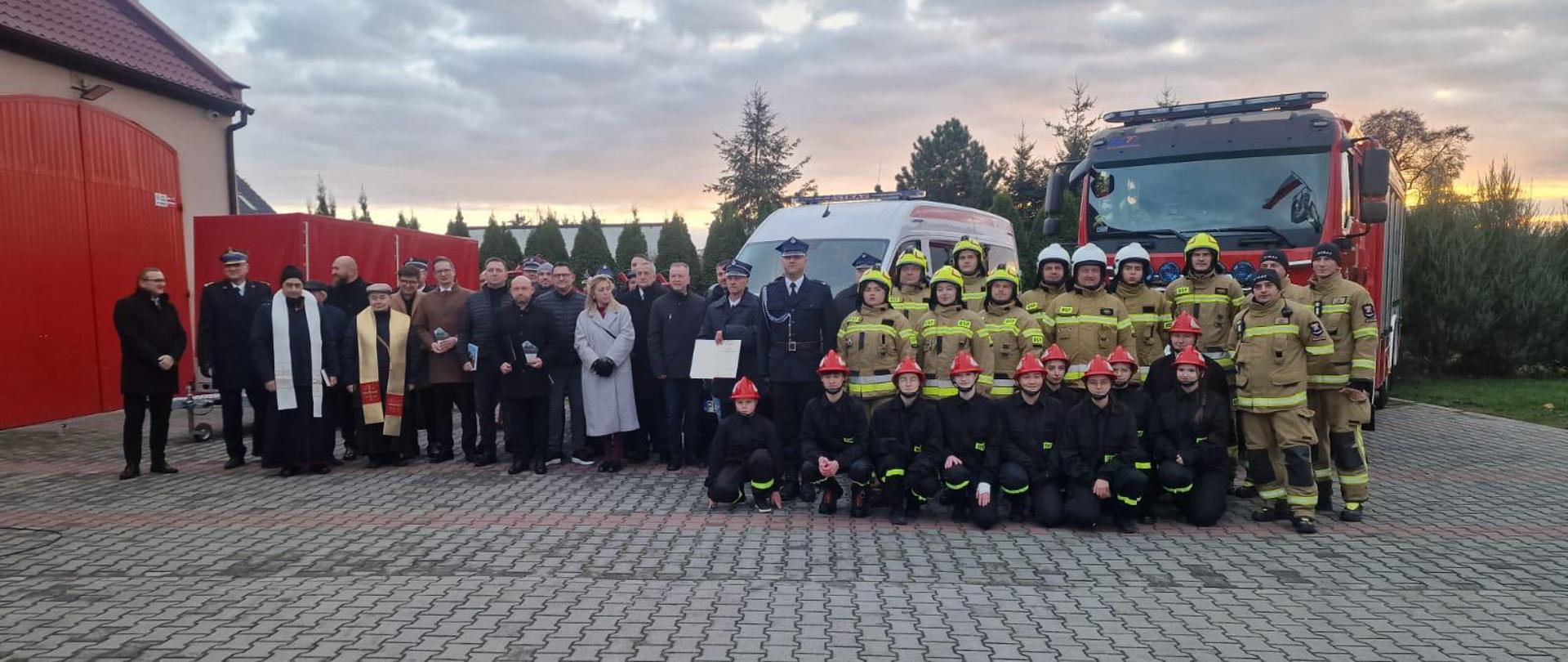 Strażacy i osoby cywilne stoją na placu w tle widoczne pojazdy strażąckie.