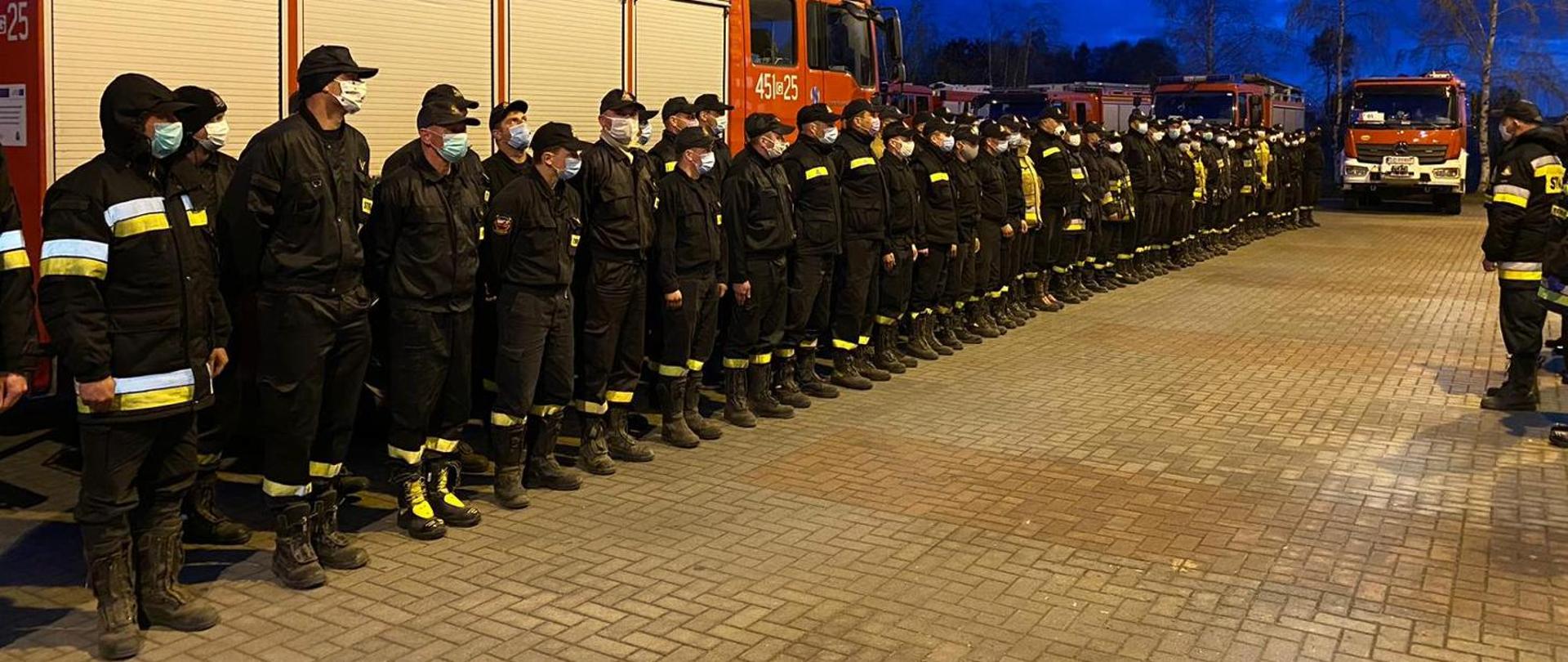 Strażacy w maseczkach ochronnych, stojący w dwuszeregu podczas zbiórki na tle samochodu. Pora wieczorowa. W oddali widoczne inne samochody pożarnicze