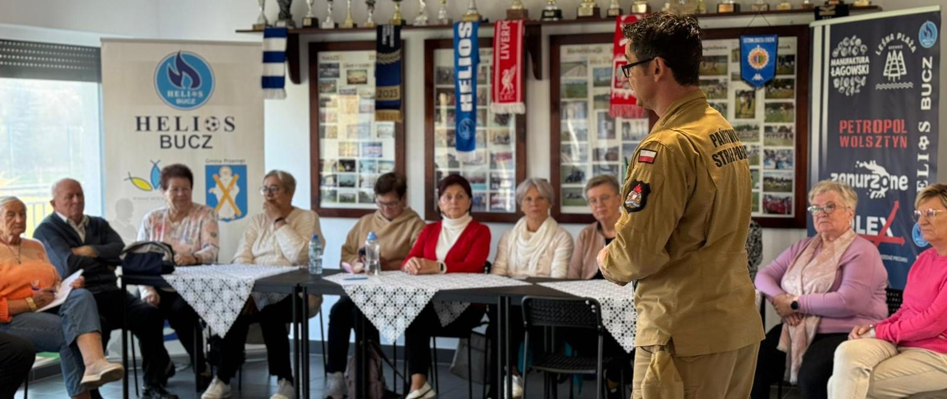 Na pierwszym planie po prawej stronie stoi bokiem strażak ubrany w piaskowe ubranie służbowe. Na drugim planie na stołem siedzą seniorzy biorący udział w spotkaniu. 