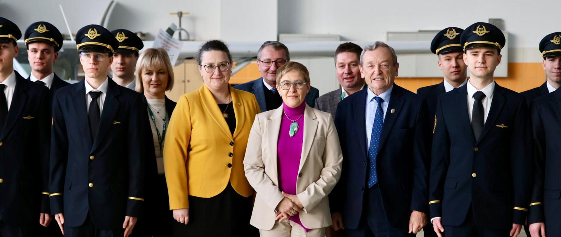 W dużej sali z białymi ścianami stoi w dwuszeregu duża grupa młodych ludzi w czarnych mundurach, pośrodku wiceminister Mrówczyńska w jasnoszarym garniturze, kobieta w żółtym sweterku i mężczyzna w garniturze.