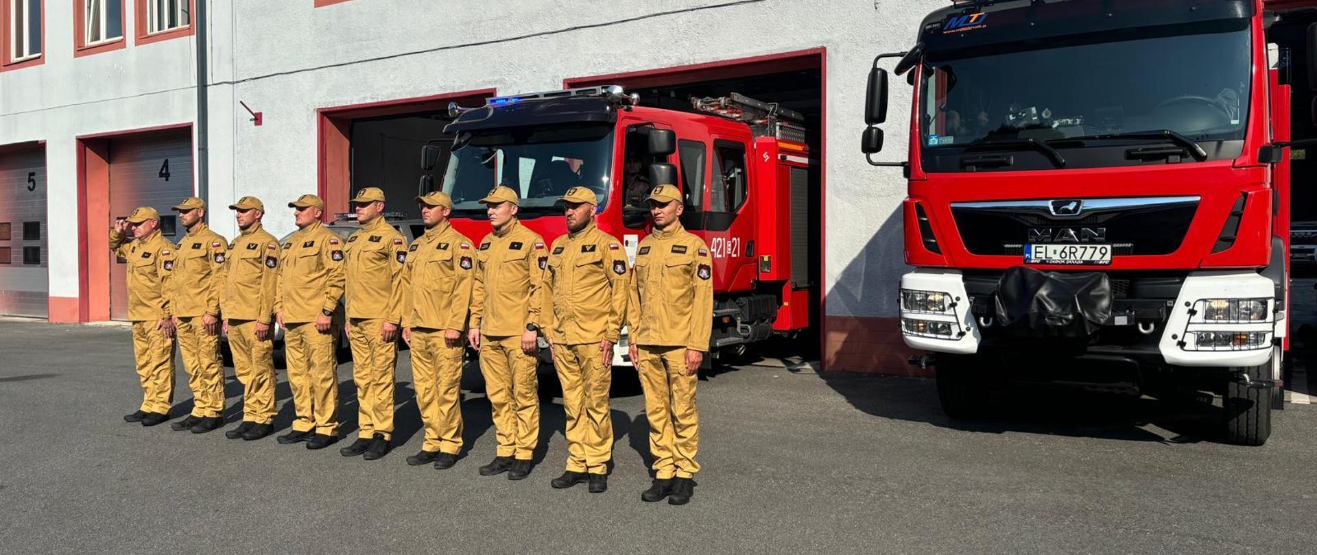 strażacy stojący w szeregu przed budynkiem jednostki, z garaży częściowo wystają samochody pożarnicze