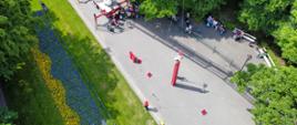Ujęcie przedstawia rozstawiony drabinę strażacką w parku. W centrum kadu widać dziecko budujące wieżę ze skrzynek. Dziecko jest asekurowane przy pomocy liny przymocowanej do kosza drabiny. Koniec liny jest trzymany przez ratownika.