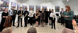 Grupa uczniów oraz pedagogów jasielskich szkół muzycznych stoi w Muzeum Regionalnym w Jaśle trzymając różnorodne instrumenty. Wszyscy są ubrani w galowe stroje. W tle widnieją fotografie. Ogólna kolorystyka czarno-beżowo-biała.