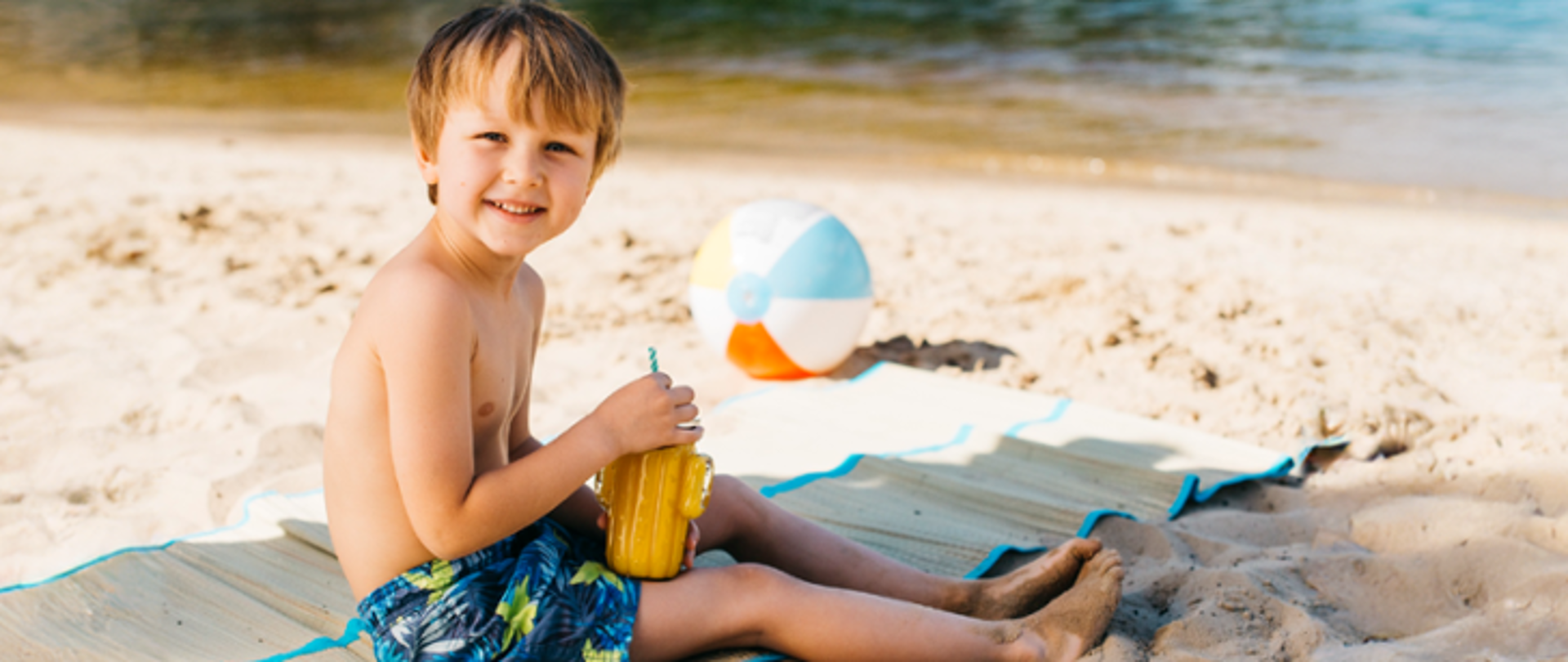Zdjęcie przedstawia chłopca na plaży, który pije sok.