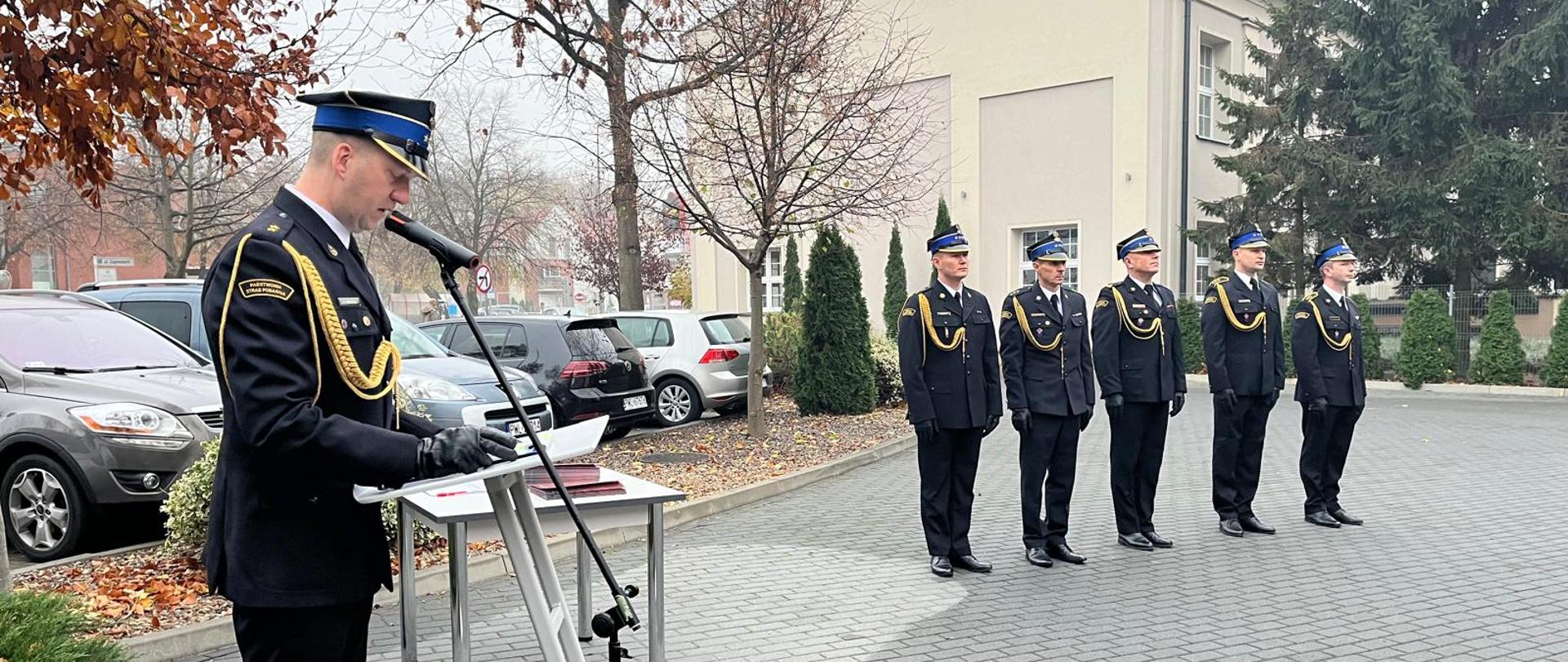 Na środku zdjęcia w umundurowaniu wyjściowym strażacy stojący w szeregu. Po lewej stronie przy mównicy i mikrofonie stoi strażak.