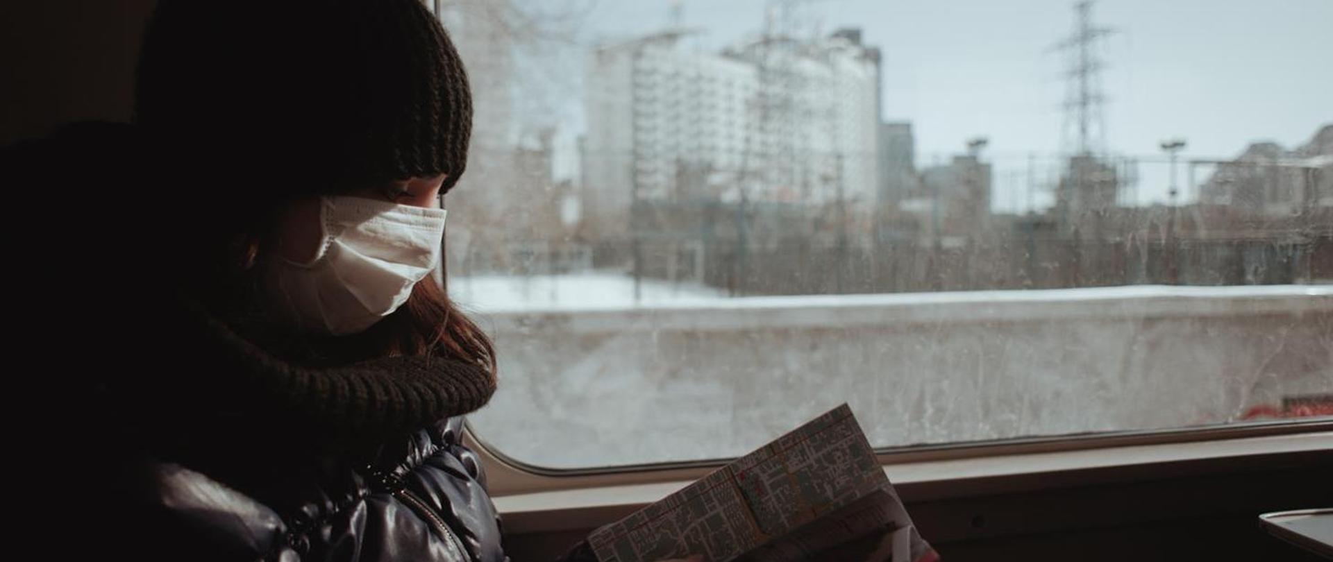 Kobieta w maseczce ochronnej na twarzy, siedząca w pociągu przy oknie, czytająca gazetę.