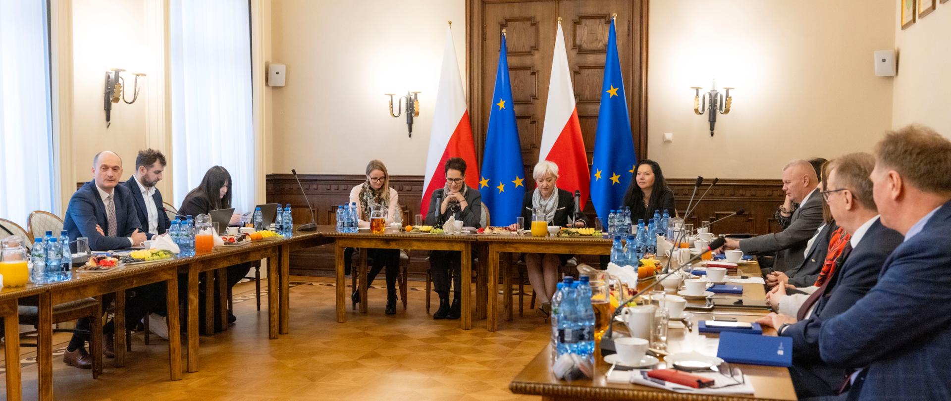 Kobiety i mężczyźni siedzą za stołem konferencyjnym ustawionym w podkowę. W tle flagi UE i Polski.