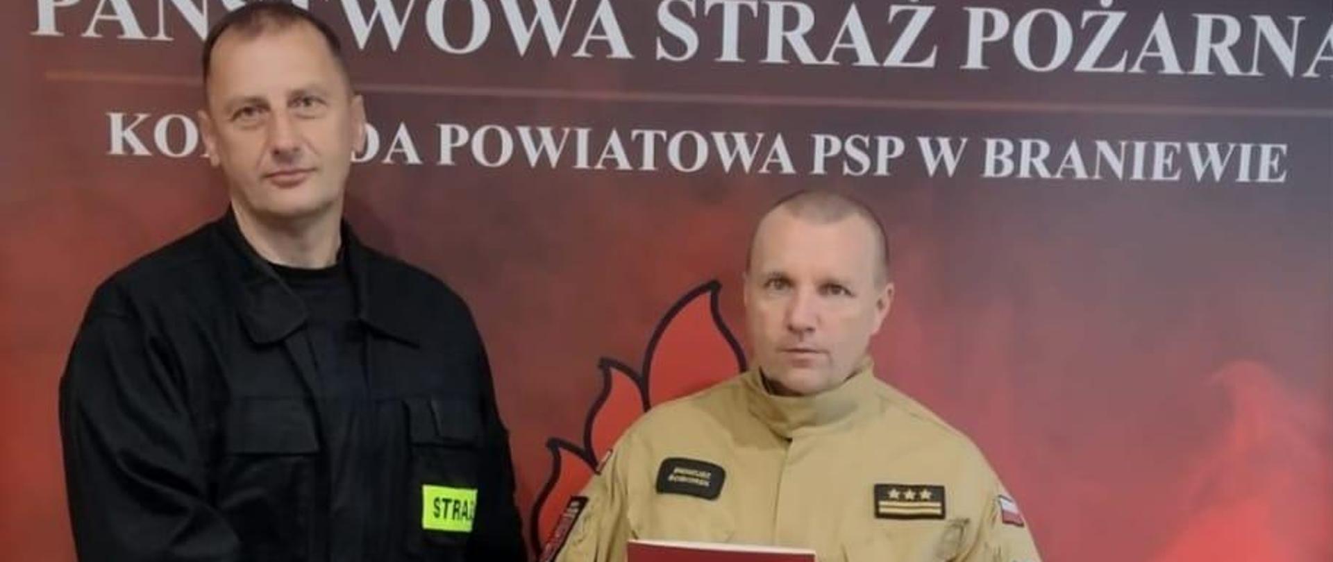 Na zdjęciu w jasnym mundurze komendant powiatowy PSP w Braniewie z lewej w czarnym mundurze strażak OSP. Stoją na czerwonym tle z napisem Państwowa Straż Pożarna.