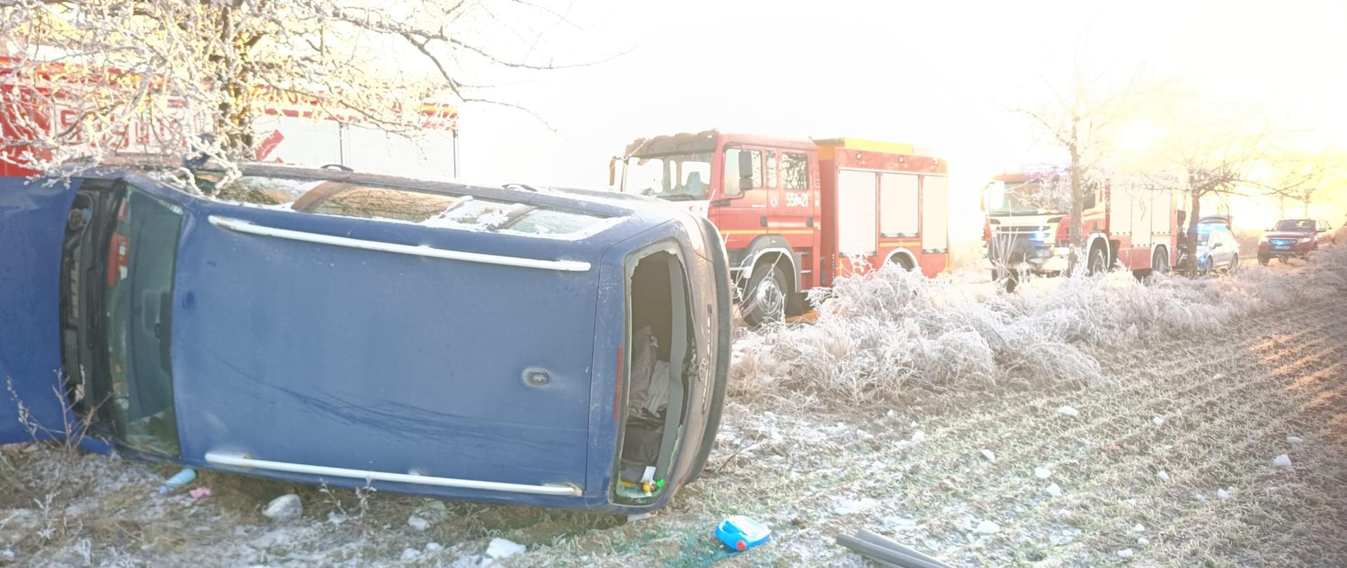 Uszkodzony samochód osobowy poza drogom, w tle wozy strażackie.