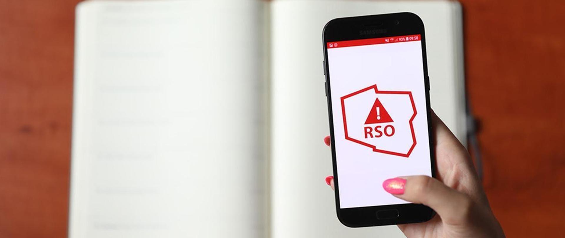 Smartfon trzymany w kobiecej dłoni. Na ekranie widoczne czerwony logotyp Regionalnego Systemu Ostrzegania na białym tle
