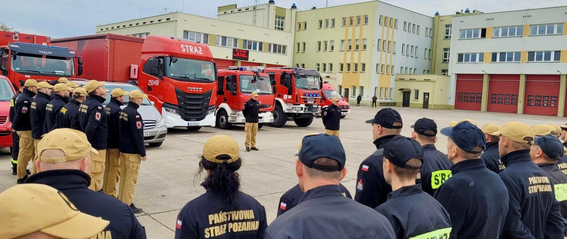 Widok na plac manewrowy przed strażnicą, gdzie trwa zbiórka strażaków. Kilkudziesięciu ratowników ustawionych w dwuszeregach, za nimi pojazdy pożarnicze, w tle budynki.