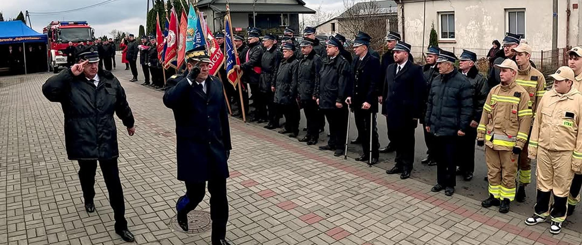 Mazowiecki Komendant Wojewódzki PSP w asyście dowódcy uroczystości dokonuje przeglądu pododdziału strażackiego. W tle poczty sztandarowe, samochód strażacki i dwa budynki mieszkalne. 