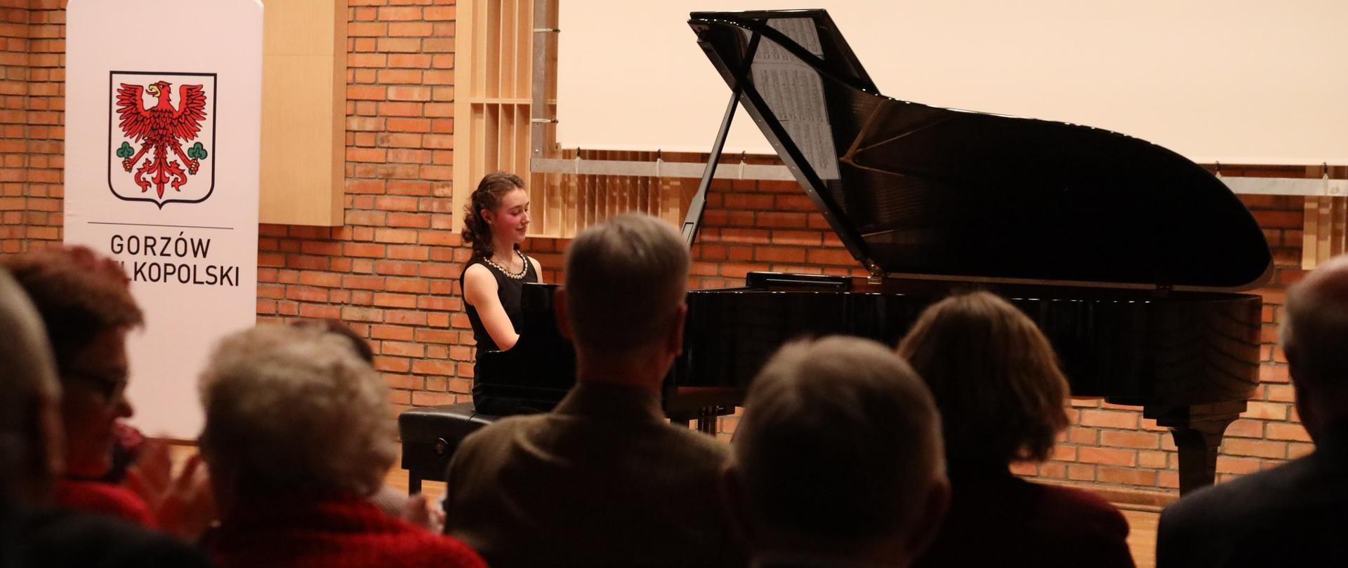 Z lewej strony logo Gorzowa. Na scenie przy fortepianie pianistka. Publiczność obserwująca koncert.