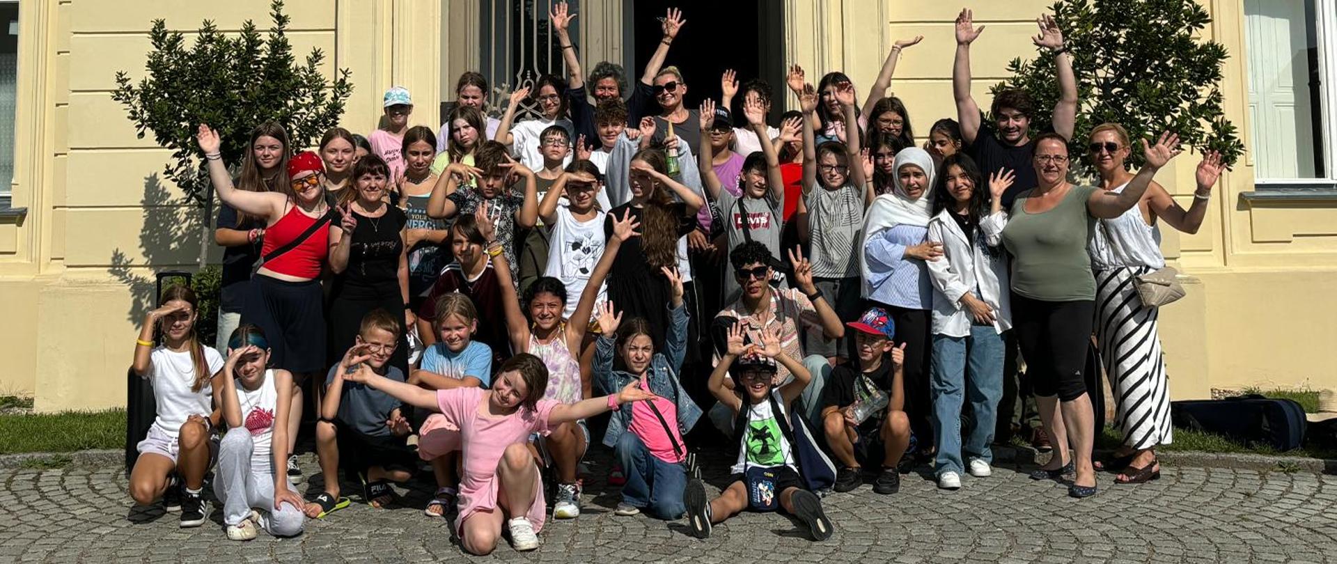 Grupa kilkudziesięciu osób, dzieci i dorosłych, stoją przed wejściem do budynku, wszyscy machają rękami do osoby fotografującej.