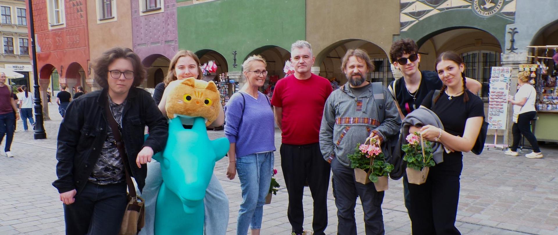 Na tle kolorowych kamienic w Poznaniu pozuje siedem osób. Jedna z dziewczyn siedzi na niebieskim dużym zwierzątku z głową przypominającą kota, inna dziewczyna i jeden z mężczyzn trzymają w rękach tekturowe koszyczki z pelargoniami, jeden z koszyczków stoi pośrodku na kostce brukowej.