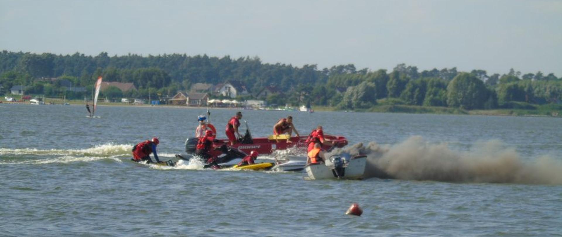 Jezioro, na wodzie czerwone i białe łodzie ratownicze, dwa skutery wodne koloru żółtego i białego. Z jednej z łodzi wydobywa się ciemny dym, powstał pożar. Ratownicy ubrani w czerwone kombinezony prowadzą działania ratownicze.