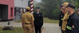 Strażacy stoją na placu przed budynkiem Komendy. Jeden z nich składa Komendantowi meldunek. Za nimi widoczny maszt flagowy z podniesioną flagą państwową. 