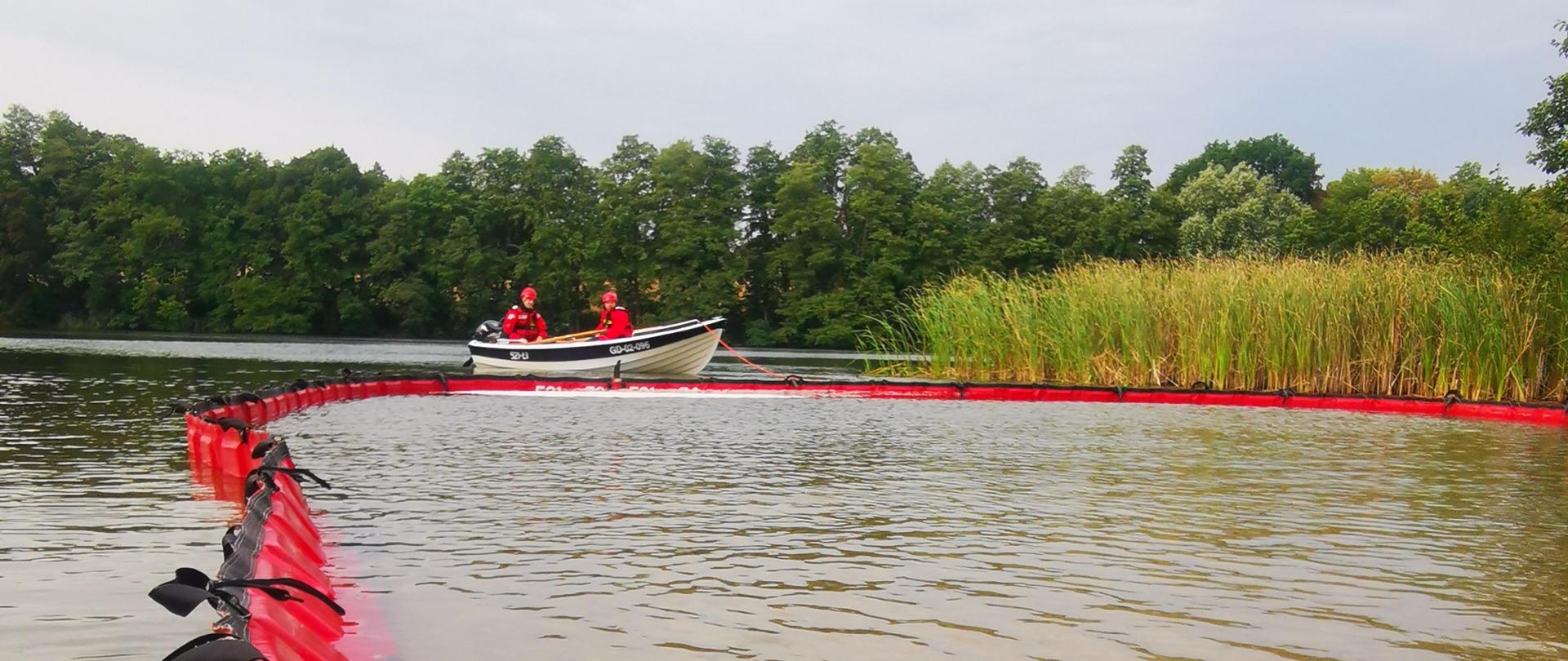 Zdjęcie przedstawia akwen wodny z rozstawioną zaporą, w tle lodź strażacka z 2 strażakami, w tle las