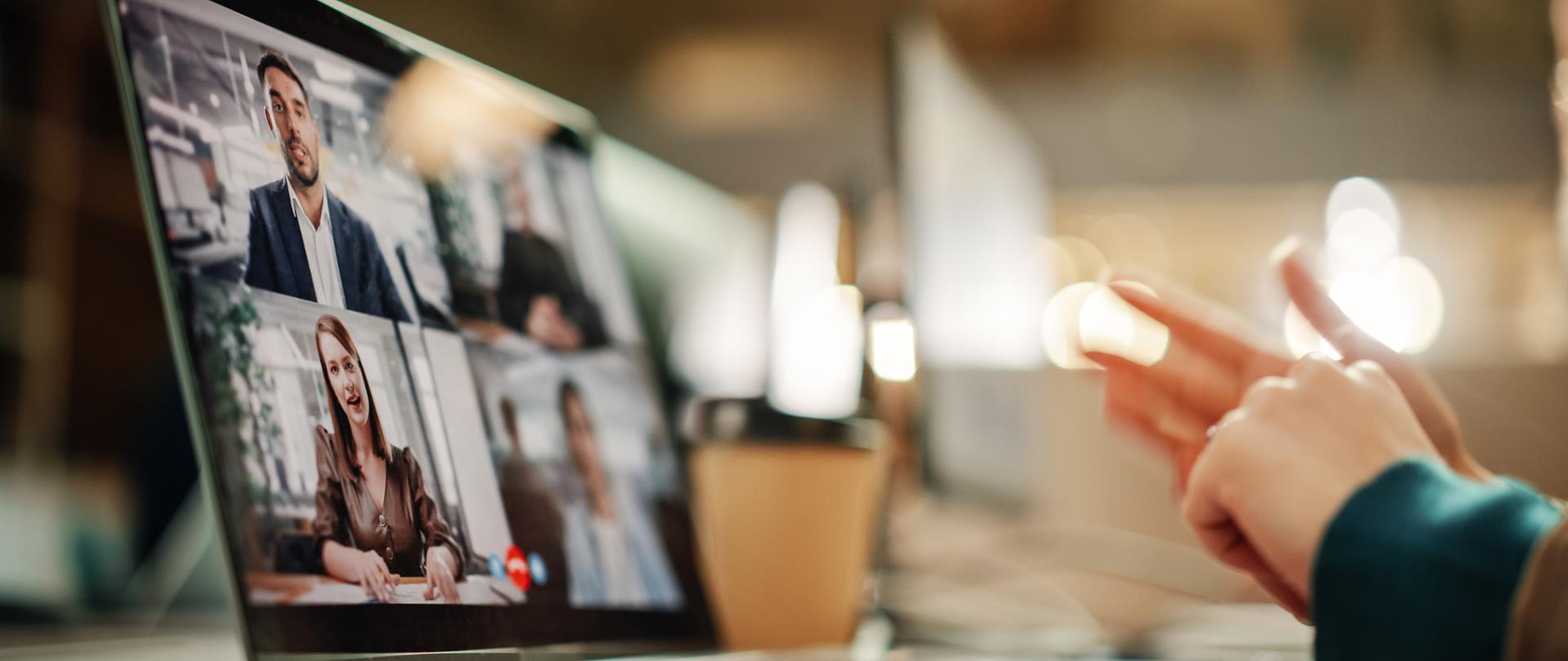 Po lewej stronie laptop, na ekranie widać pulpit spotkania online i okienka z uczestnikami, po prawej ręce osoby siedzącej przy biurku