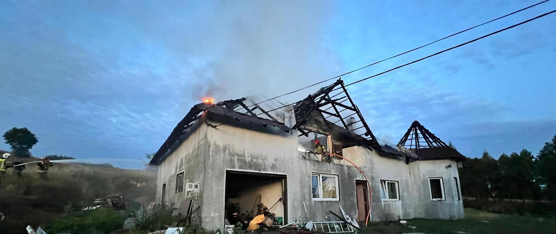 Na zdjęciu widoczny dom mieszkalny oraz strażak dogaszający dach budynku