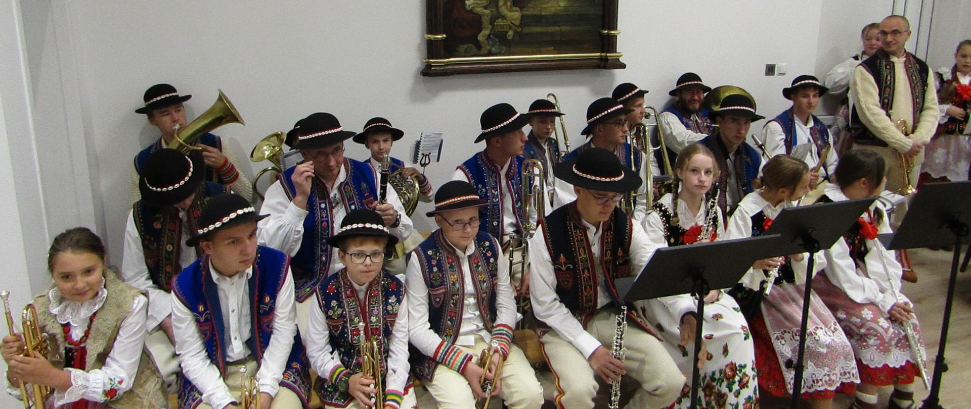 Aula przykościelna, młodzież siedzi w strojach góralskich w rękach trzymają instrumenty, przed nimi pulpity za nimi na ścianie płachta z ikona świętej. 