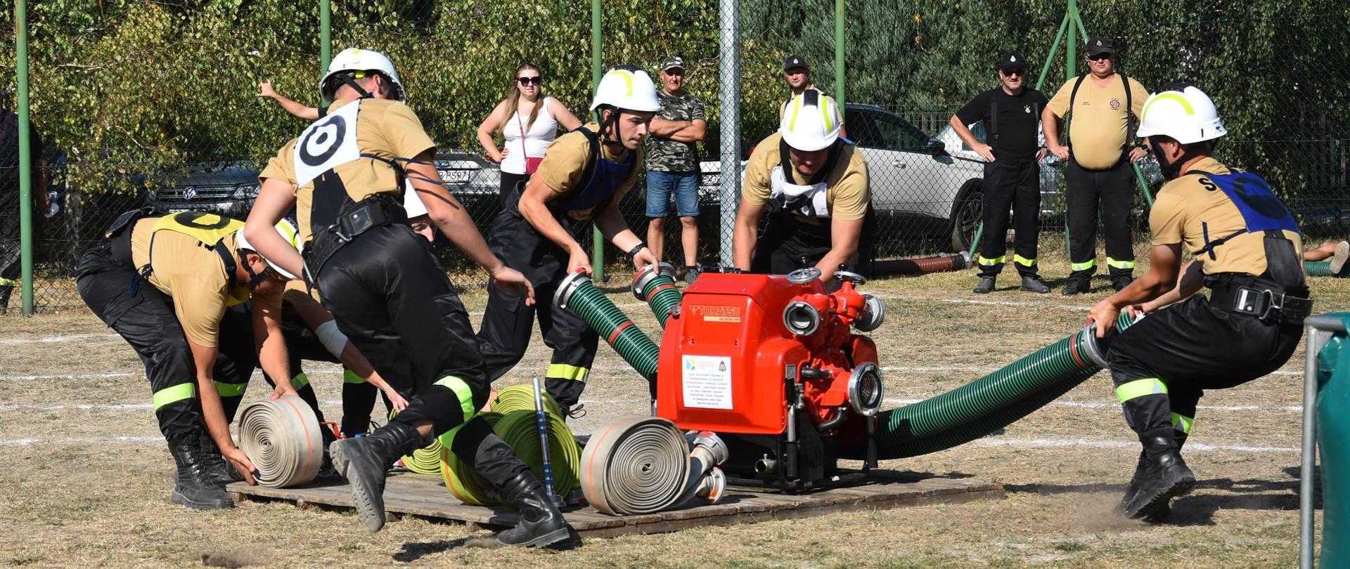 Boisko piłkarskie, sześciu strażaków OSP w ubraniu koszarowym oraz w koszulkach piaskowych z krótkim rękawkiem w białych hełmach, po wystartowaniu dynamicznie sprawiają armaturę wodną, jeden druh odpala motopompę pożarniczą ustawioną na podeście drewnianym. W tle kibice przyglądający się wykonywaniu ćwiczenia.