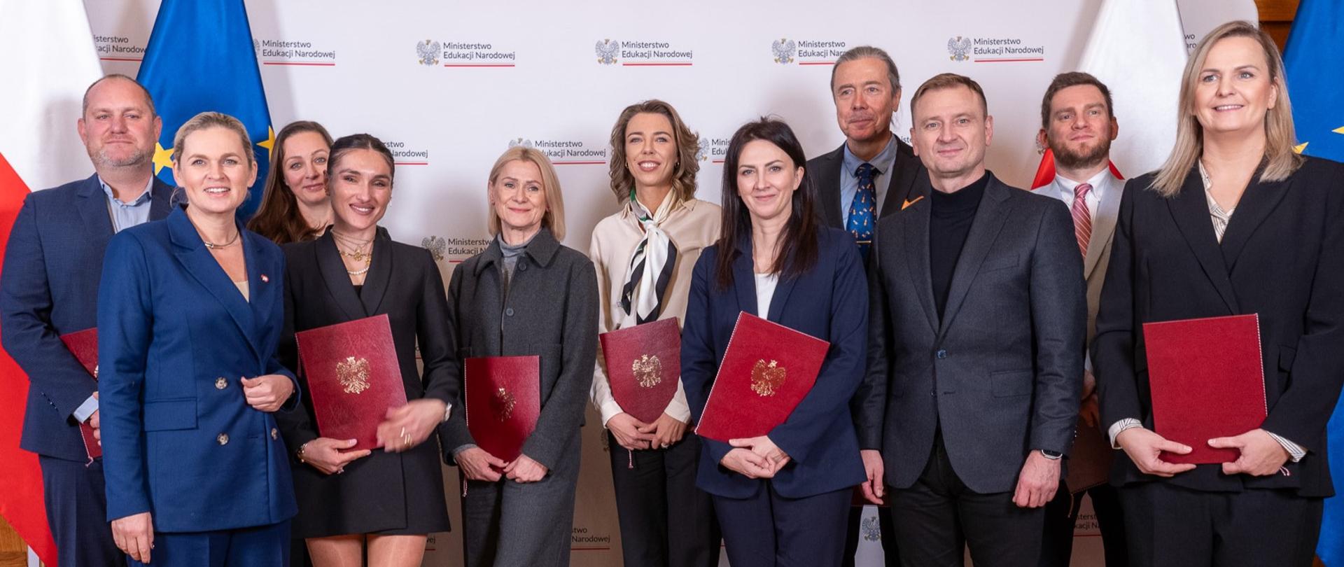 Zdjęcie zbiorowe Na tle białej ścianki z napisem Ministerstwo Edukacji Narodowej stoi grupa ludzi, którzy w rękach trzymają bordowe teczki złotym godłem wśród nich minister Barbara Nowacka i minister Sławomir Nitras 