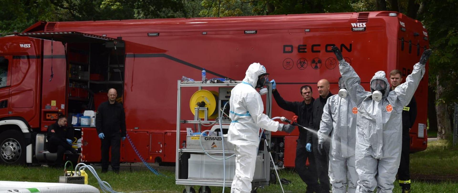 Samochód do działań CRNE stoi na otartym terenie, przed nim stoi kilku strażaków, 3 z nich w skafandrach do działań chemicznych, jeden spłukuje wodą z węża skafander drugiej osoby