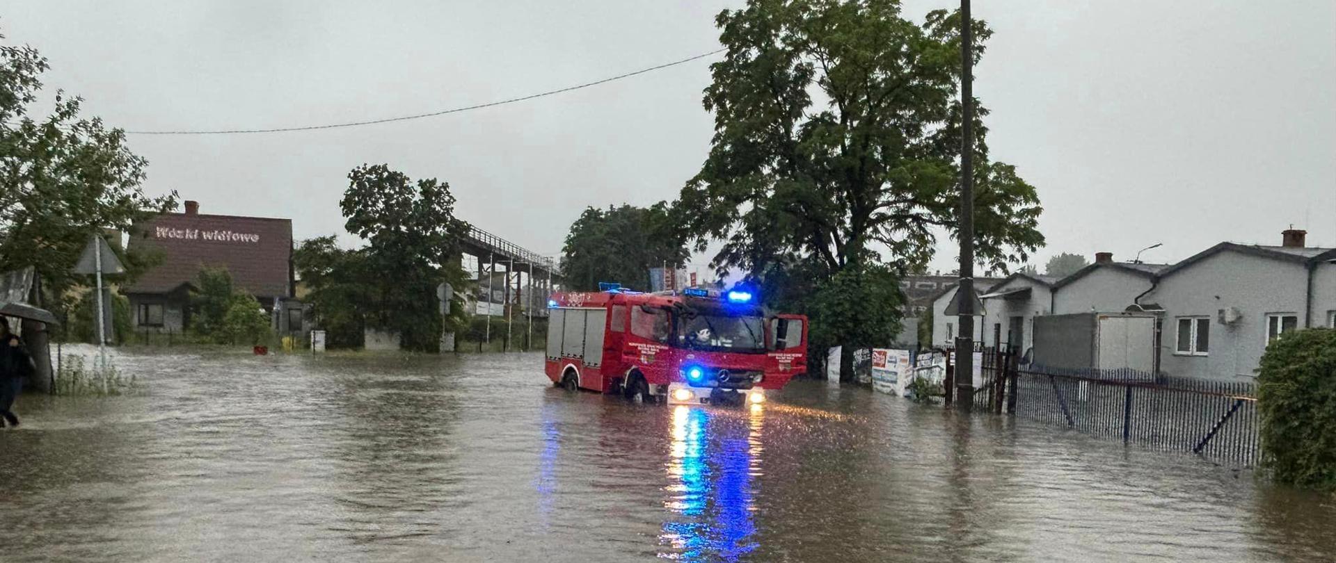 Wóz strażacki stoi zaparkowany na środku zalanej ulicy, ma włączone niebieskie sygnały świetlne. 