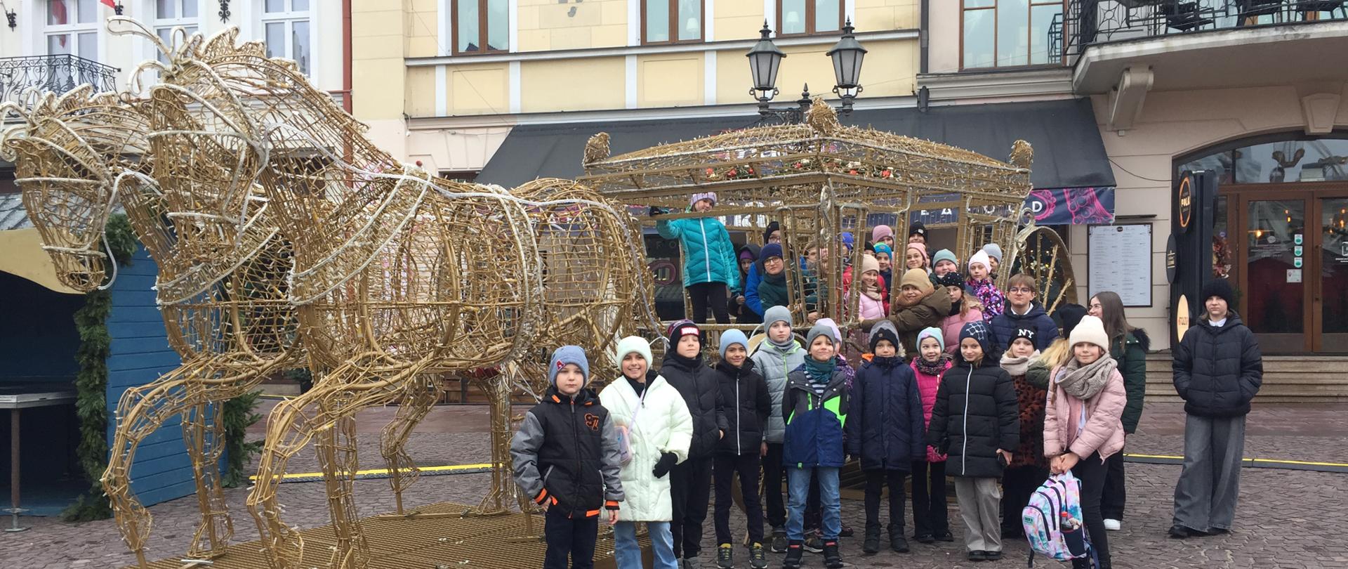 Uczestnicy wycieczki stoją na rynku w Rzeszowie przed konstrukcją metalową tworzącą karocę oraz ciągnące je konie. Zdjęcie jest robione w ciągu dnia, dzieci są w kurtkach zimowych i czapkach. W tle zabudowania rzeszowskiego rynku.