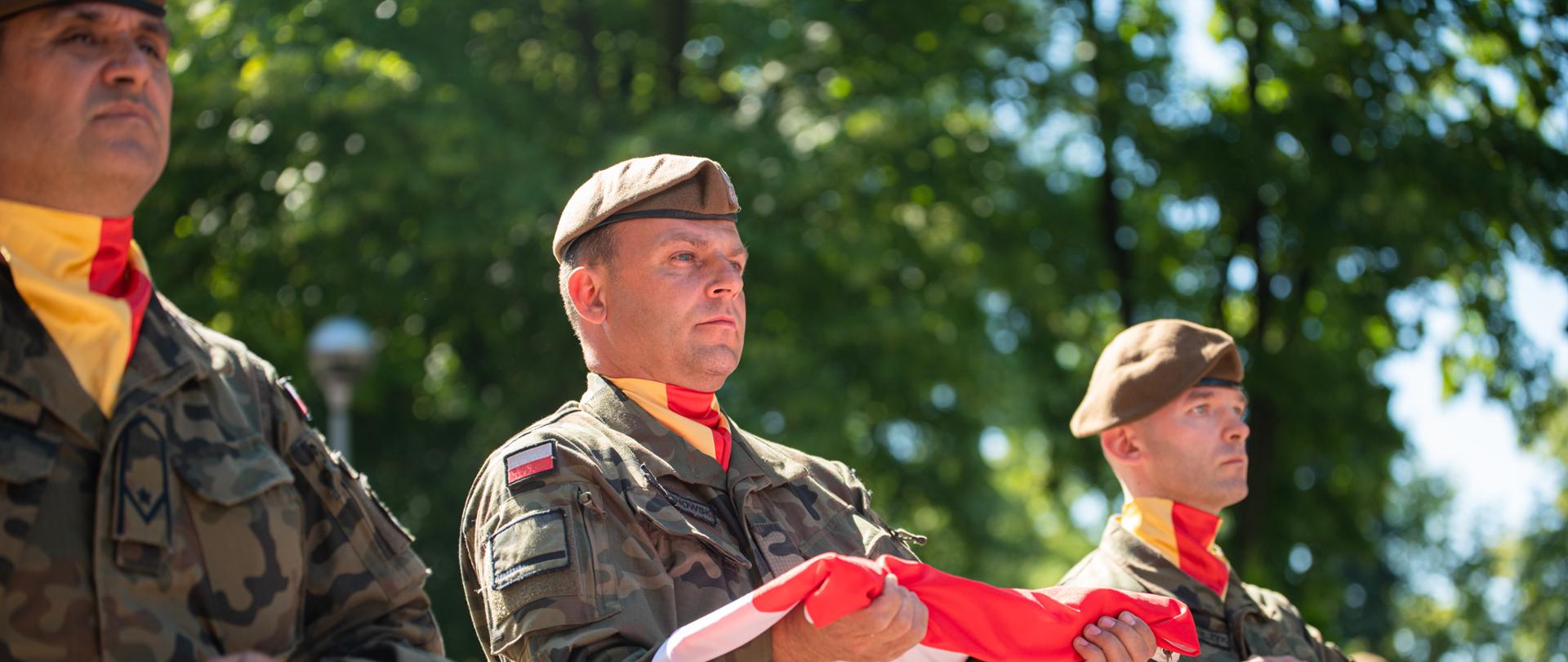 trzech żołnierzy WOT maszeruje w rzędzie, żołnierz w środku trzyma złożoną flagę Polski