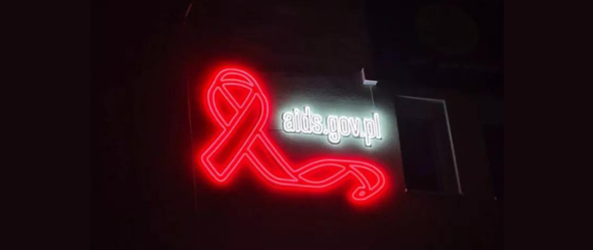 Na zdjęciu znajduje się neon z napisem aids.gov.pl w kolorze białym. Po lewej stronie znajduje się czerwona wstążka. Tło jest czarne.