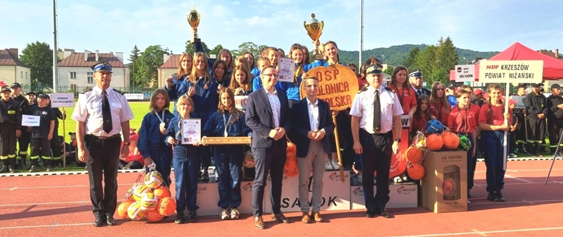 Kolorowa fotografia wykonana na zewnątrz w pogodny dzień na stadionie sportowym. Przestawia drużyny młodzieżowe na podium z nagrodami, pucharami, medalami i tablicami z nazwami jednostek. Przed drużynami stoją oficerowie Państwowej Straży Pożarnej i osoby cywilne.