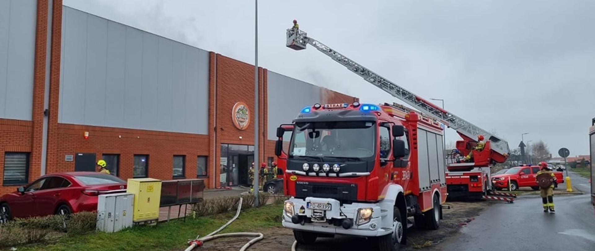 Samochód strażacki stoi na drodze obok na ziemi porozwijane węże strażackie prowadzące do budynku. Obok stoi strażacka drabina mechaniczna i strażacy.