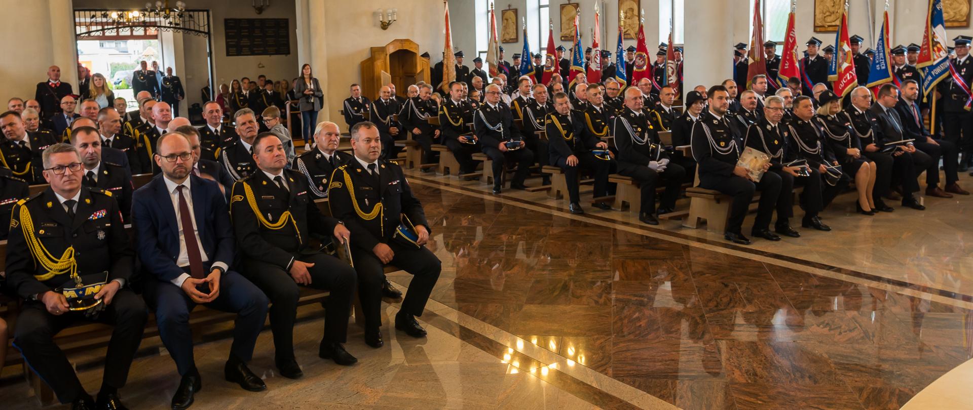 strażacy osp siedzący w ławkach w kościele, za nimi w nawie bocznej poczty sztandarowe, zdjęcie wykonane w przodu z ukosa