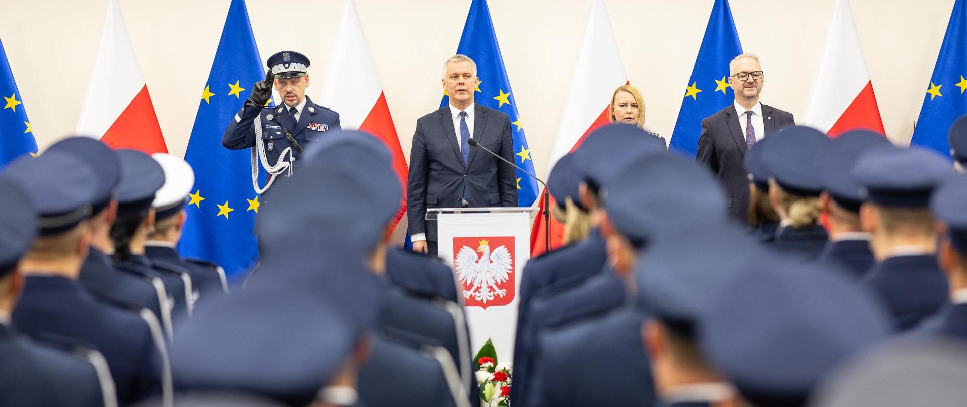 Minister Siemoniak stoi przy mównicy. Obok niego komendant Boroń i rektor-komendant Malasińska-Nagórny. Na pierwszym planie stojący plecami przyszli oficerowie Policji 