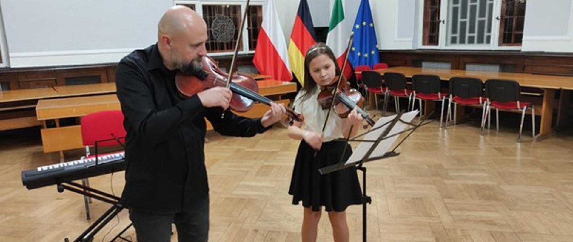 Zdjęcie kolorowe. Milena ze skrzypcami ubrana w białą bluzkę i czarną spódnicę oraz nauczyciel Artur Pastuszyński ze skrzypcami ubrany na czarno.