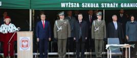 Na dworze w słońcu pod zielonym namiotowym daszkiem z napisem Wyższa Szkoła Straży Granicznej stoi grupa ludzi, z boku mównica obok której stoi kobieta w ceremonialnym czerwonym stroju z gronostajowym kołnierzem.