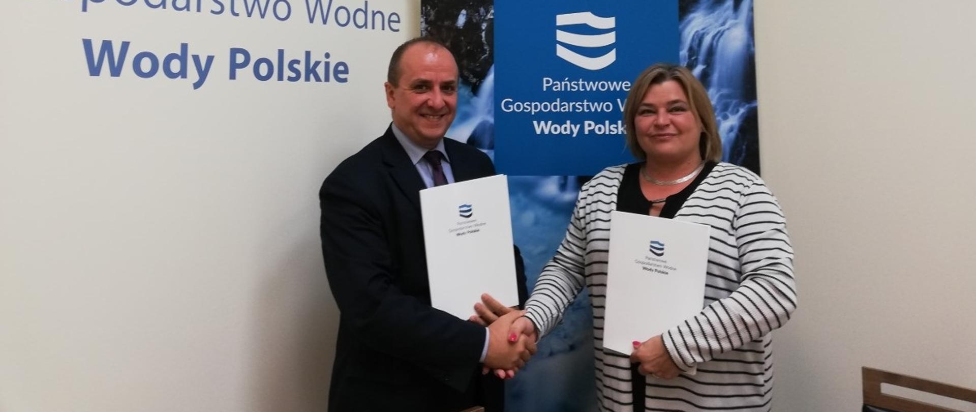 Pani Agnieszka Szymula i Pan Profesor Radosław Dobrowolski na tle plakatu z logotypem Wód Polskich. Ściskają sobie dłonie, trzymają białe teczki.